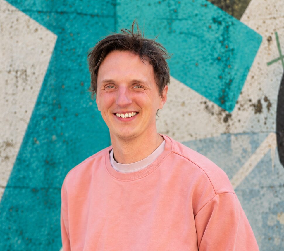 James smiling, wearing a salmon coloured jumper. Against a green and white geometric background.