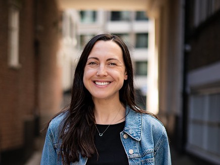 Genevieve smiling against background of building with windows. Wearing black top and denim jacket.