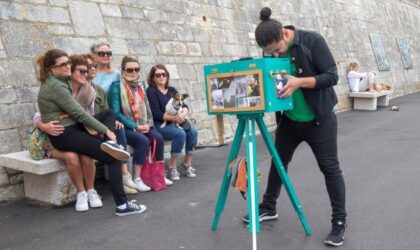 Events To the left, five women sit on a stone bench against a stone wall. One of them holds a dog on their lap. They are watching a man who is bent over and looking into a re imagining of an old fashioned light-box camera on a tripod. The camera is a sea green. Then man wears his hair in a bun.