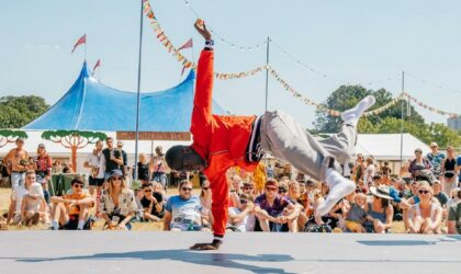 Events A man wearing a red jacket and cream trousers break-dances on a low stage in front of an audience who are seated on the grass. There is a blue sky and a festival tent in the back ground.