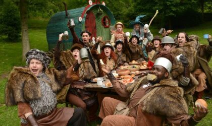 Events A group of people sit outside and around a long table. They are dressed like Hobbits. They are laughing and raising their glasses in a toast.
