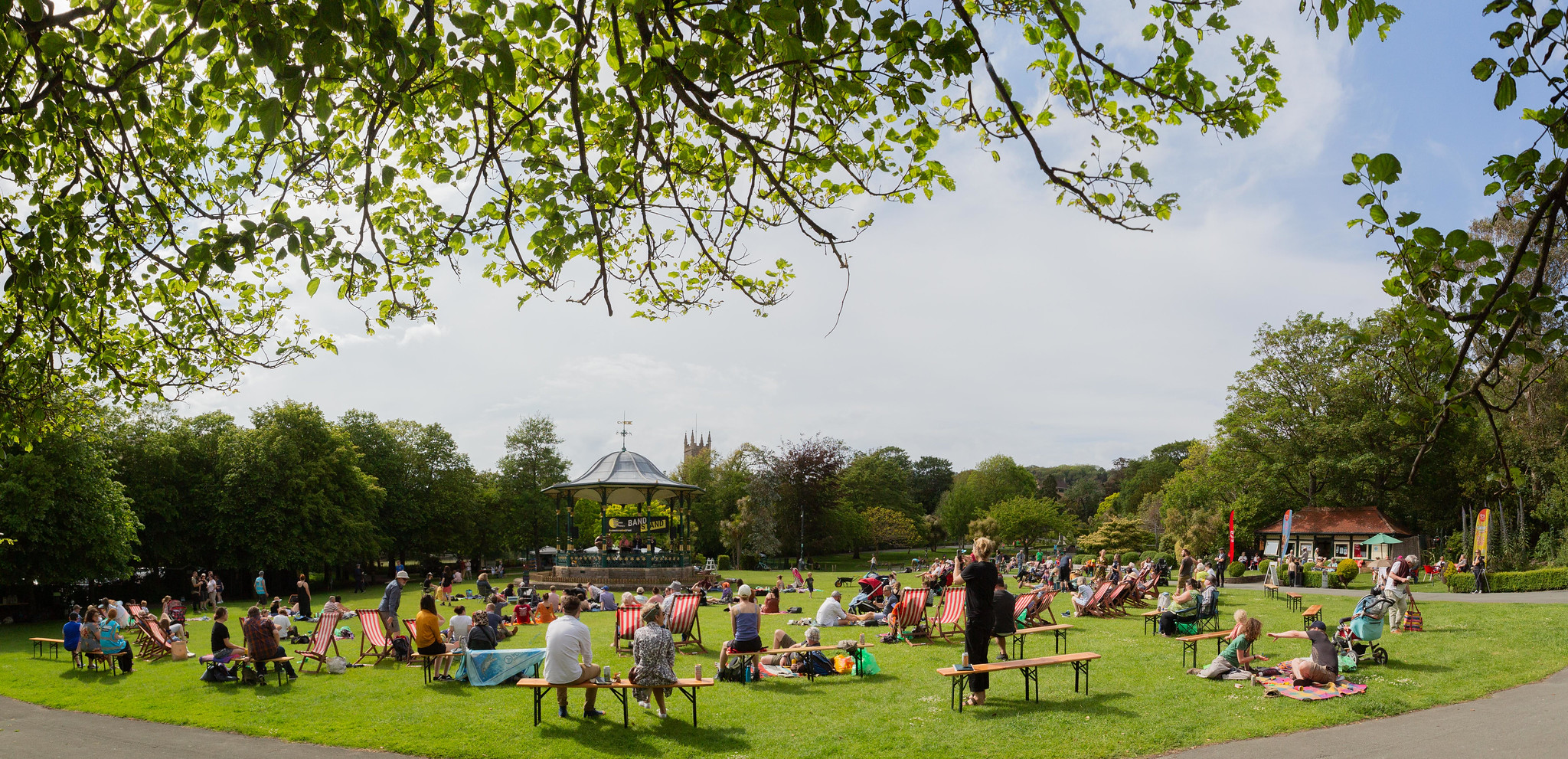 A park scene with a bandstand in the background and spectators in the foreground sitting on the floor, benches and deck chairs