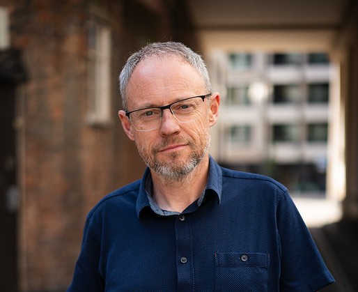 Andy wearing a blue shirt and glasses, against a backdrop of a brick wall and windows.