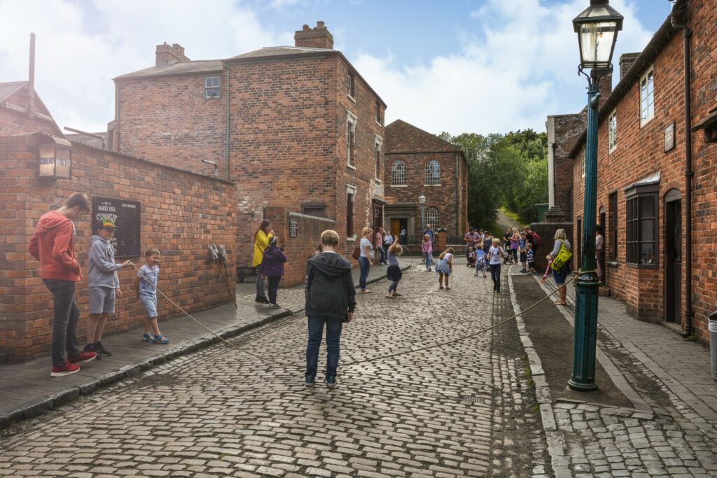 A cobbled street is lined by red brick houses. A child wearing a cap stands on the left side of the street holding a skipping rope. The other end is tied to a victorian era lampost on the right side of the street. A child in a hoodie is about to jump over the rope. There is a small crowd of children and adults in the distance.