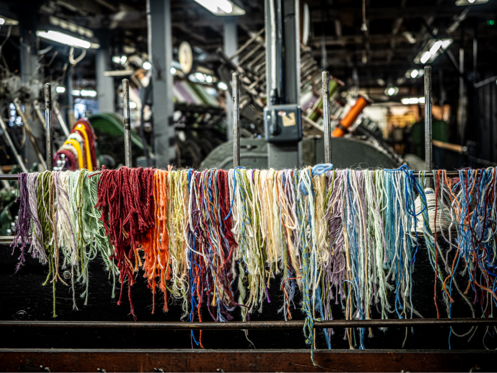 A wool production factory. Large metal machines dominate the background. In the forefront, hundreds of colour wool scraps and hung over a thin pipe creating a rainbow of wool.