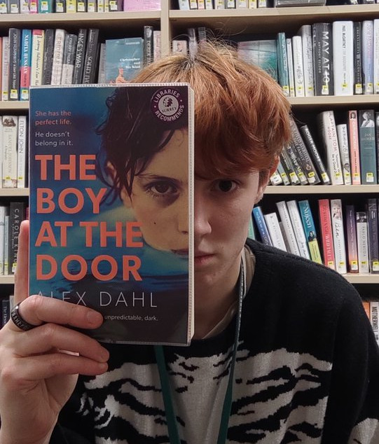 A young person in a library. They hold a book up to their face, and the book cover features a half face. The book is positioned over half of their face, giving the illusion that the book and person are one and the same.