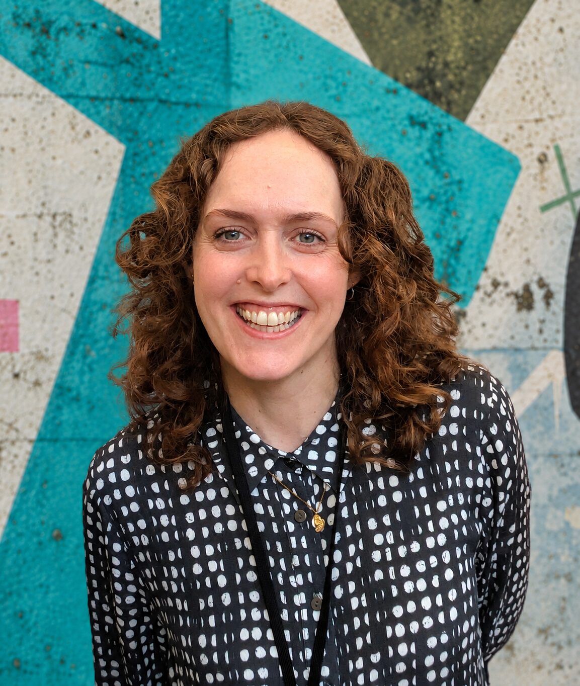 Julia smiling, wearing a black blouse with white spots. Against a green and white abstract background.