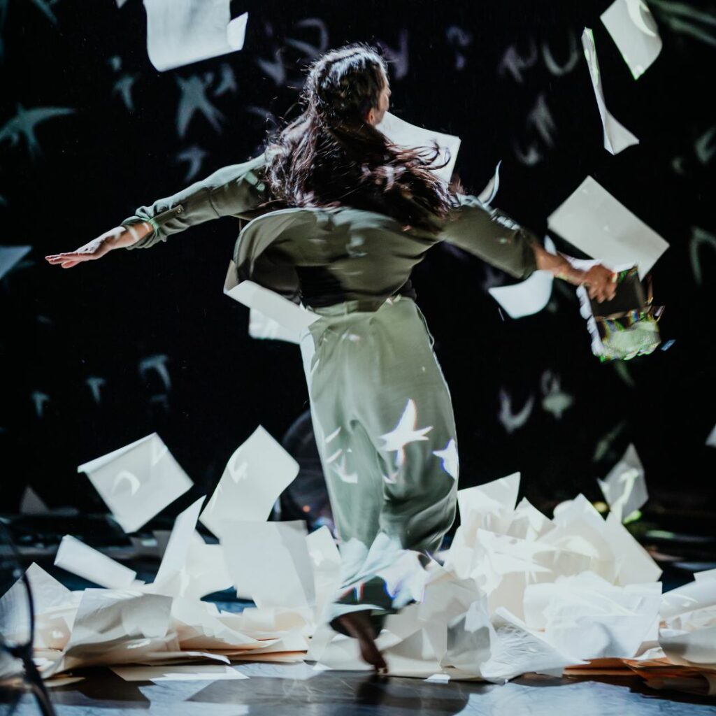 A woman is swirling with her arms outstretched. She is dancing amongst a swirl of papers.