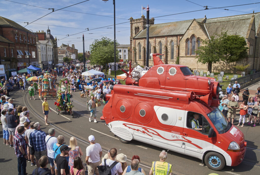 An ariel view of hundreds of festival goers lining a street. They watch a parade of performers, led by a red van and white vehicle which has been altered to resemble a spaceship.
