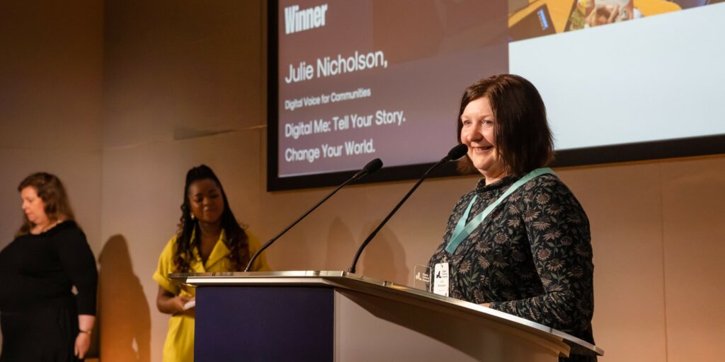Julie, a woman with short dark hair wearing a dark floral dress, is accepting her award on stage. She smiles as she speaks into lectern microphones. Beyond Julie is Emma-Louise, the host, who is wearing a bright yellow jumpsuit, Beyond Emma-Louise is the BSL interpreter, who is signing for the audience. 