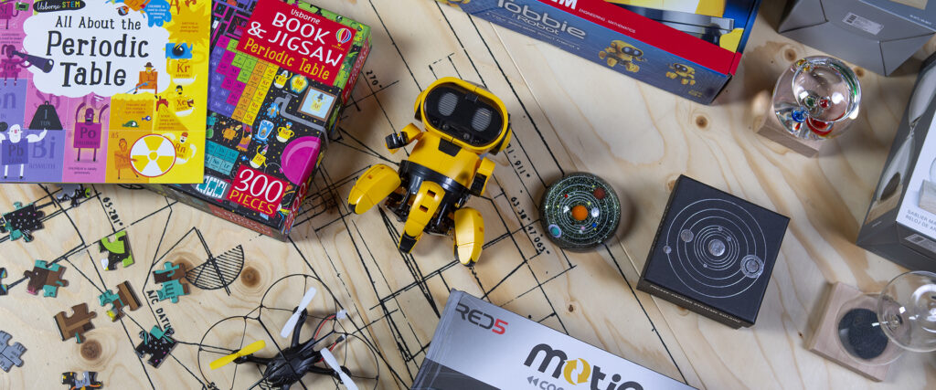 Pictured are products from the We The Curious website, as part of a flatlay. Products include a book and a jigsaw puzzle about the periodic table, a yellow robot, a mini drone and small models of the solar system. The lay upon a pale wooden board. 