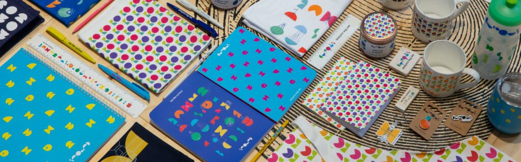 Pictured are products from the We The Curious website, as part of a flatlay. Products include colourful branded notebpads, pens, mugs, candles, badges, aprons, rulers, waterbottles and teatowels.