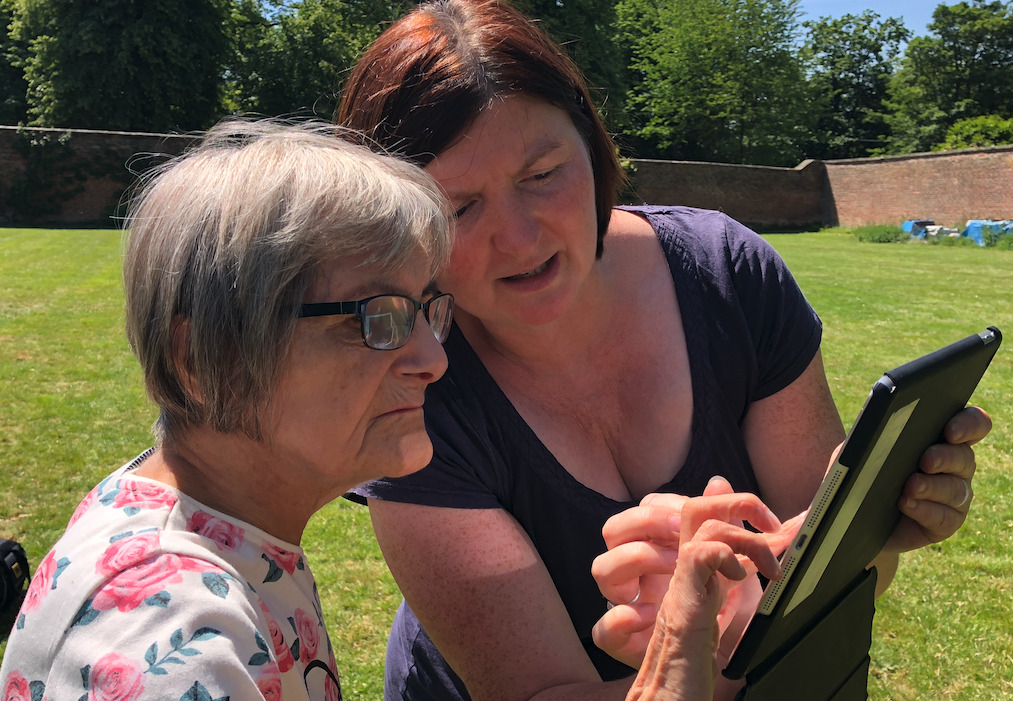 It's a sunny day and two women are in a large lawned area. One has grey hair and glasses, and the other brown hair wearing a blue t-shirt. Both are interacting with a digital tablet.