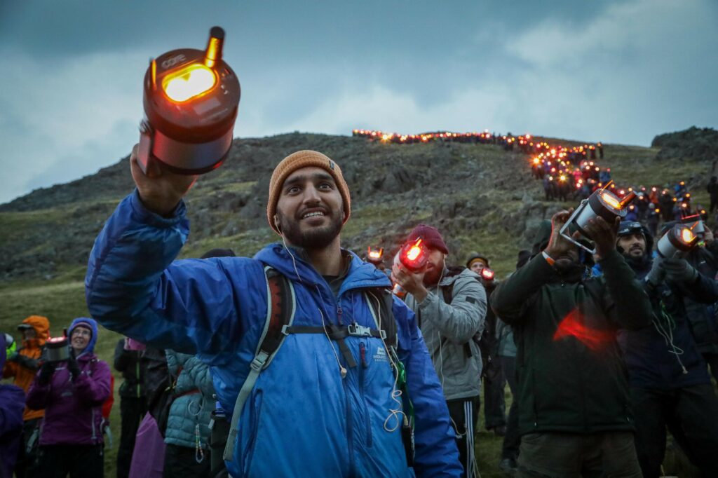 A member of the group Muslim Hikers holding a geolight at the top of Scafell Pike. Behind him hundreds of people hold their geolights snaking back up to the top of the mountain.