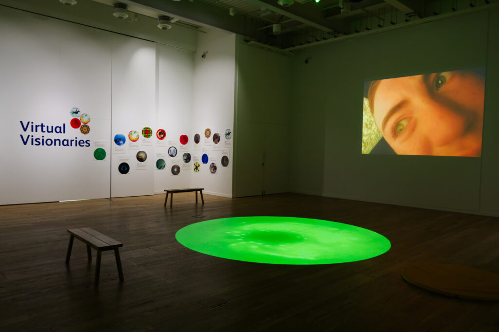 A gallery space with white walls and wooden floors. The back wall features the words 'Virtual Visionaries' and a collection of colourful, round art works. There is a large green circle projected onto the floor of the cemtre of the room, and two benches situated next to the circle. The wall to the right shows a projection of a person in close up. 