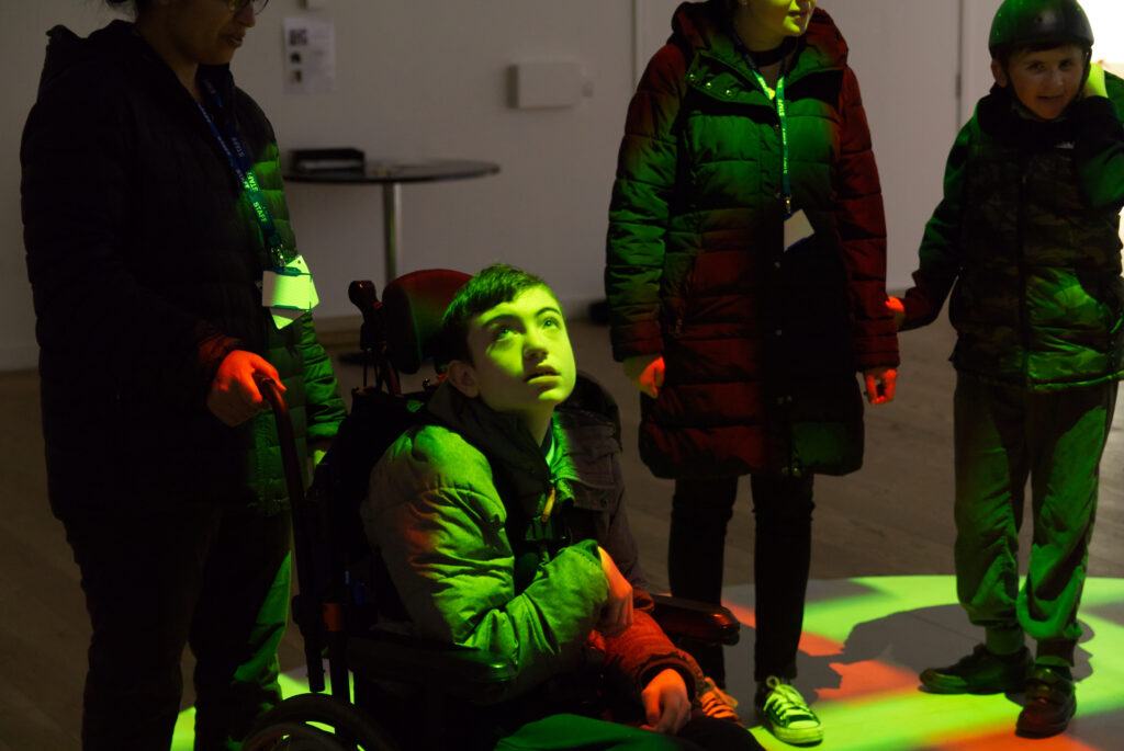 A young wheelchair user is bathed in green and red light, as they experience the Virtual Visionaries exhibition.