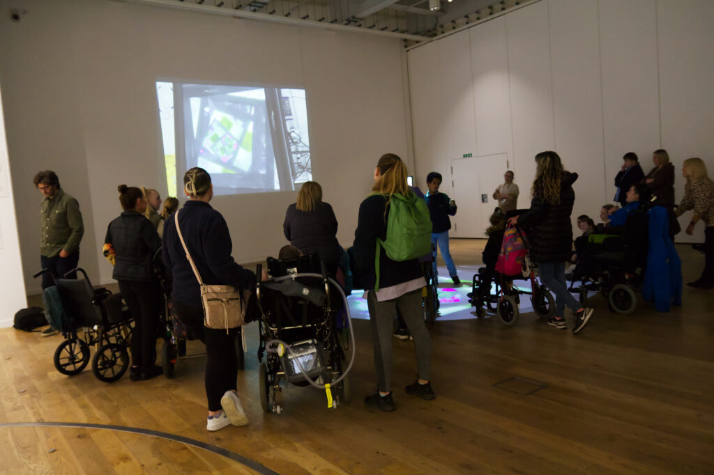 Students, teachers and parent carers are in a large studio room with white walls and wooden floors. Some are watching an image project on a screen, whilst others interact with projections on the ground beneath them.