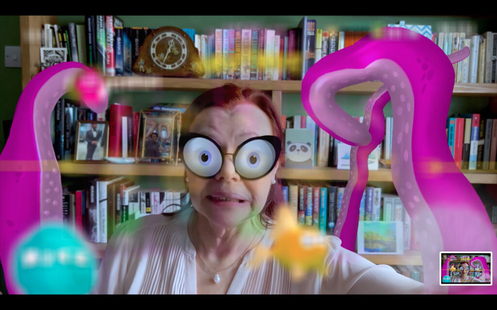 A women with red hair wearingpale pink top sits infront of a bookshelf. She is morphing into an animated creature with wide eyes and bright pink tentacles. She looks in some distress.