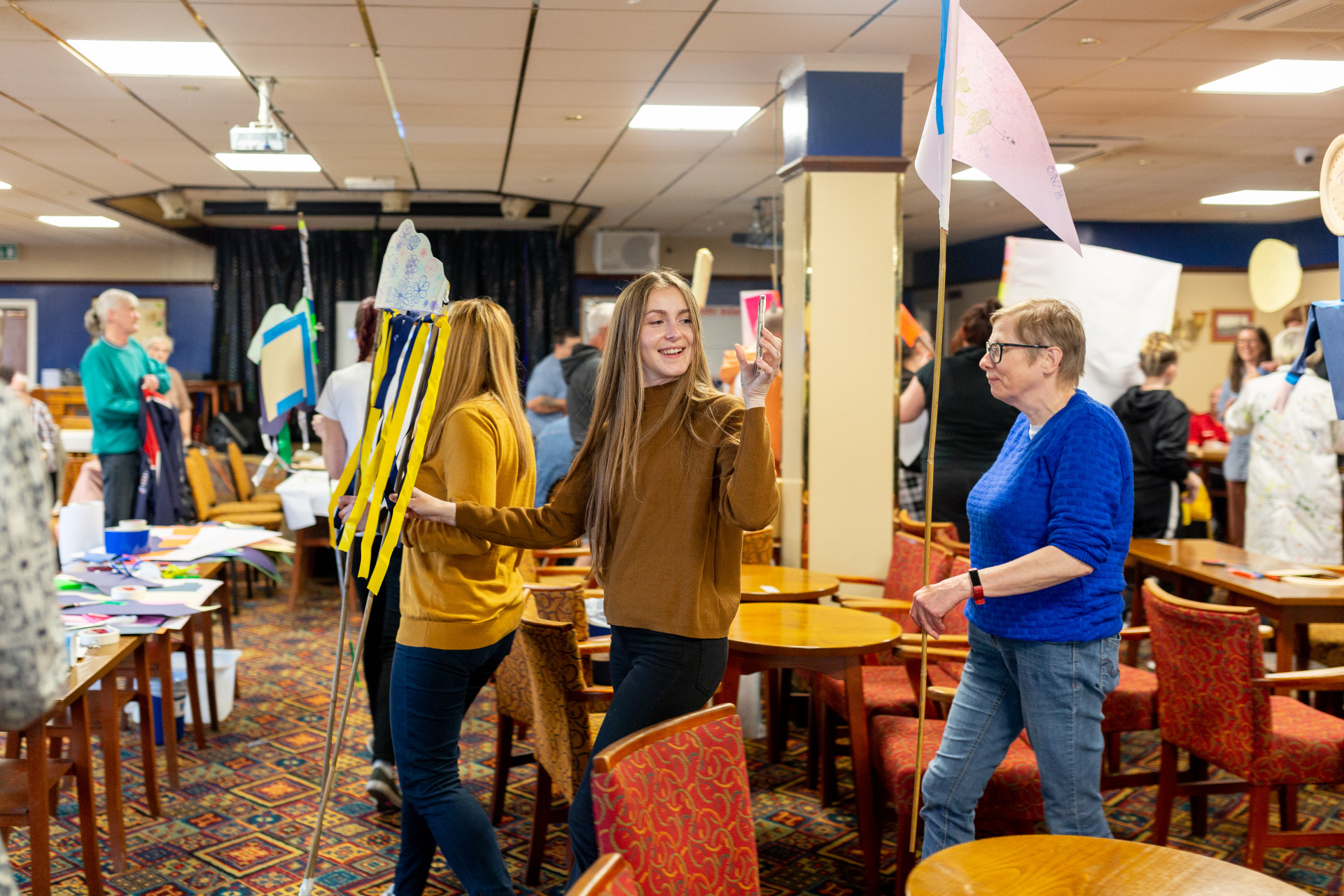 A group of people are walking around a large room with colourful carpet and lots of tables a chairs. Many of them are holding handmade flags on long sticks. A women in the forefront with long brown hair is smiling and taking a photo of the action on her phone.