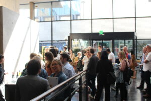 A group of people mingling in the foyer of a building. There are big windows letting in a lot of natural light and a concrete wall to the left with a drinks table in front. In the foreground there is a women wearing a denim top holding a laptop talk to a man with brown hair wearing a grey suit jacket.