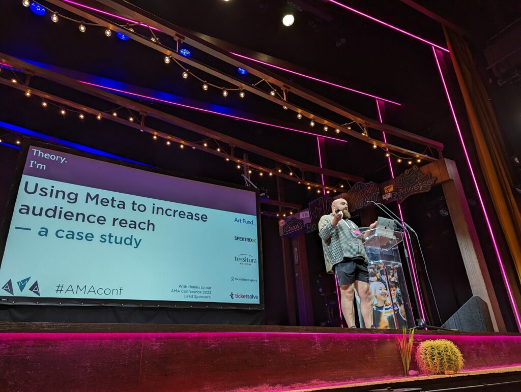 A man on stage in front to a big screen with a presentation showing.