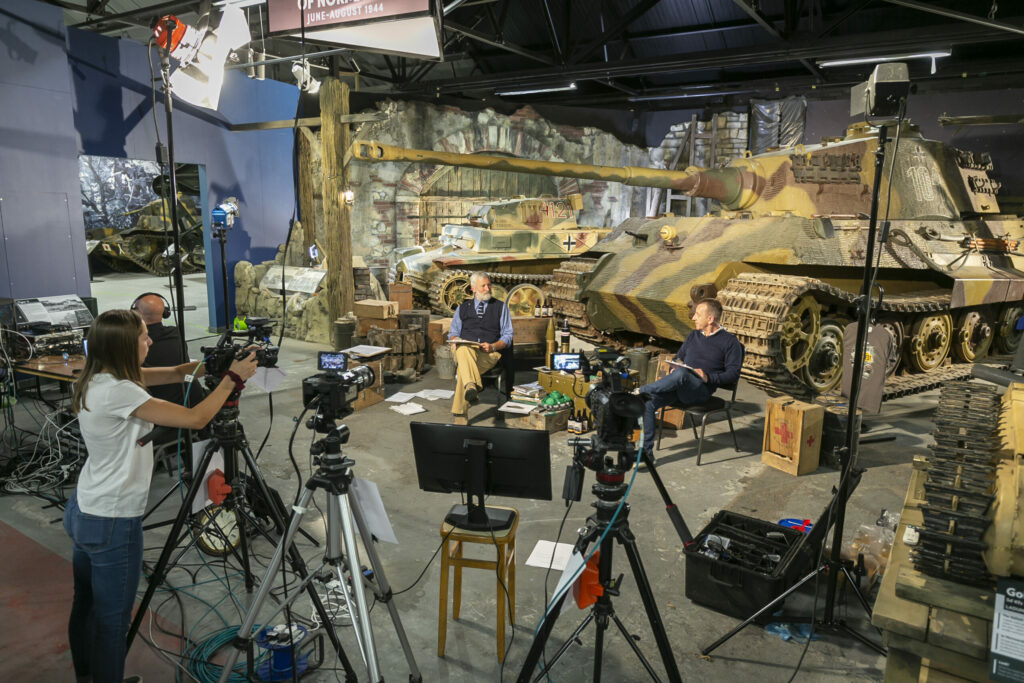 The image shows a filming studio with lots of cameras and two people sat in front of a large tank.