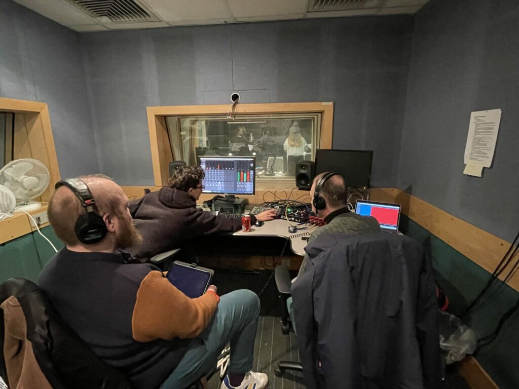 Three members of the creative team wearing headphones sit in a small recording studio, centered around a sound desk and monitor.