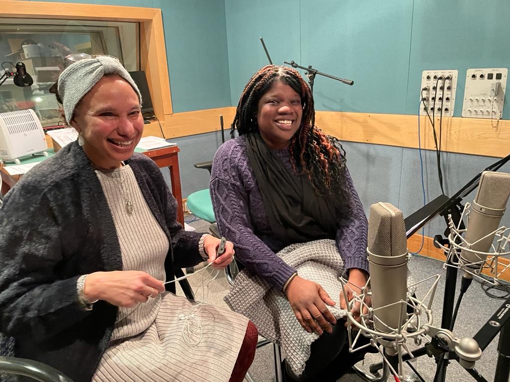 Two visually impaired voice actors sit in front of microphones in a small recording studio, smiling broadly.