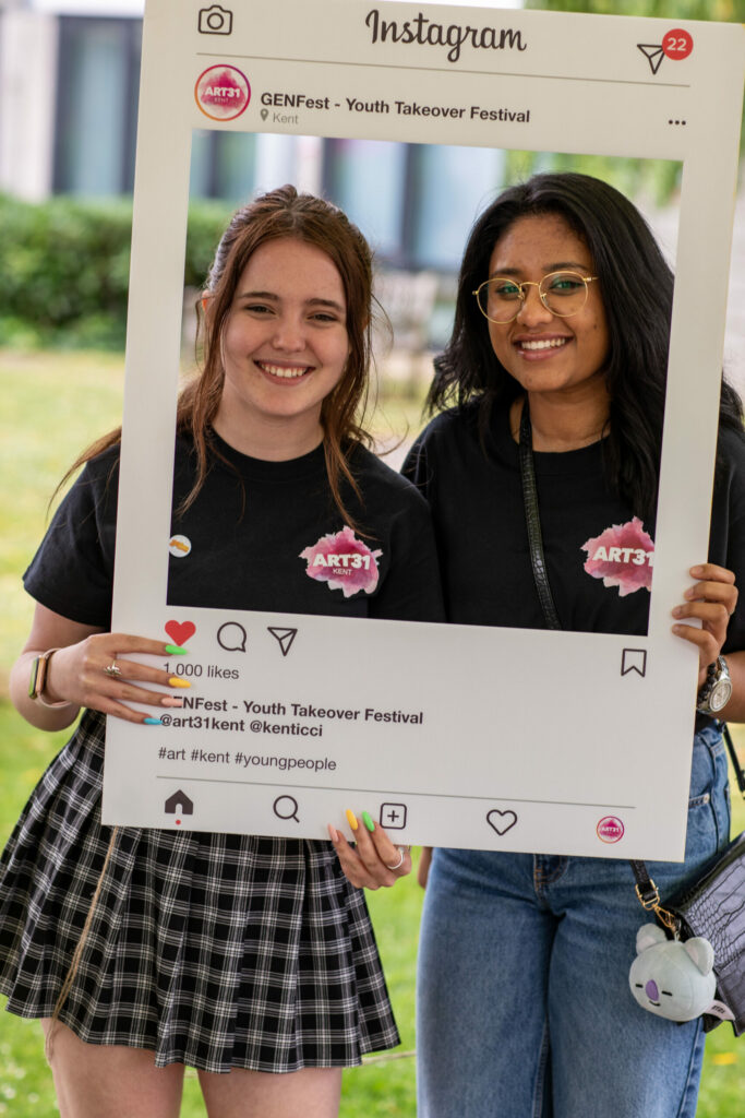 Two girls standing outside, holding a large cut out of an Instagram frame in front of them, and smiling.