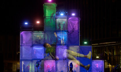 Events A multi-coloured sculpture made of different boxes with people inside them. And a person projected onto them.