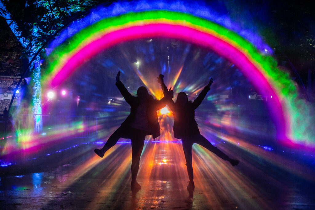 Two silhouetted people can be seen standing in starfish positions underneath an artificially lit rainbow.
