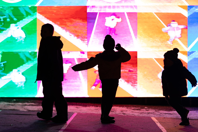 Two silhouetted children standing facing different directions on a background made up of green, red, pink and orange square digital screens.