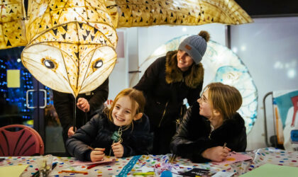 Two young girls and a woman are smiling at a paper illuminated owl to the left of them. The two girls are also doing crafts on a table whilst the woman is stood up behind them.