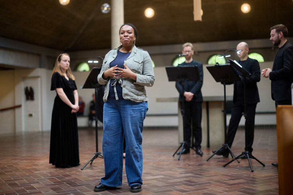 A woman wearing blue jeans and a khaki shirt performs with a consort dressed in black inside of a historical music venue.